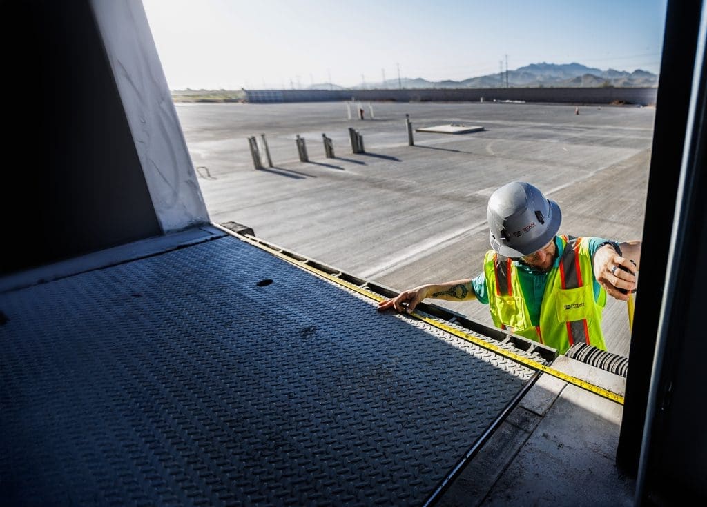 A construction worker in a safety vest and helmet measures a loading dock opening at an industrial site, demonstrating the importance of 24/7 Emergency Overhead Door Repair, with an empty parking lot in the background.