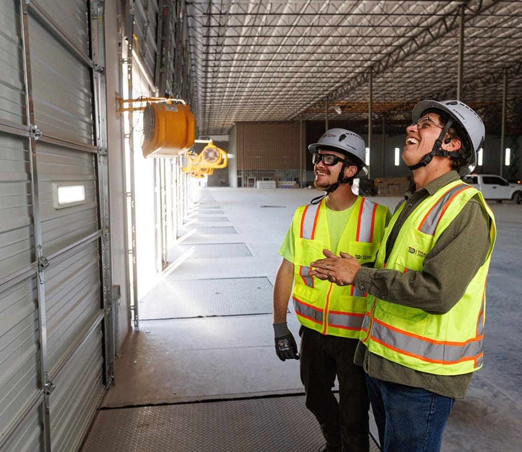 Two construction workers wearing safety gear and reflective vests stand inside a large industrial building, looking up and gesturing toward the ceiling—showcasing the expertise of Arizona Commercial Door Company.