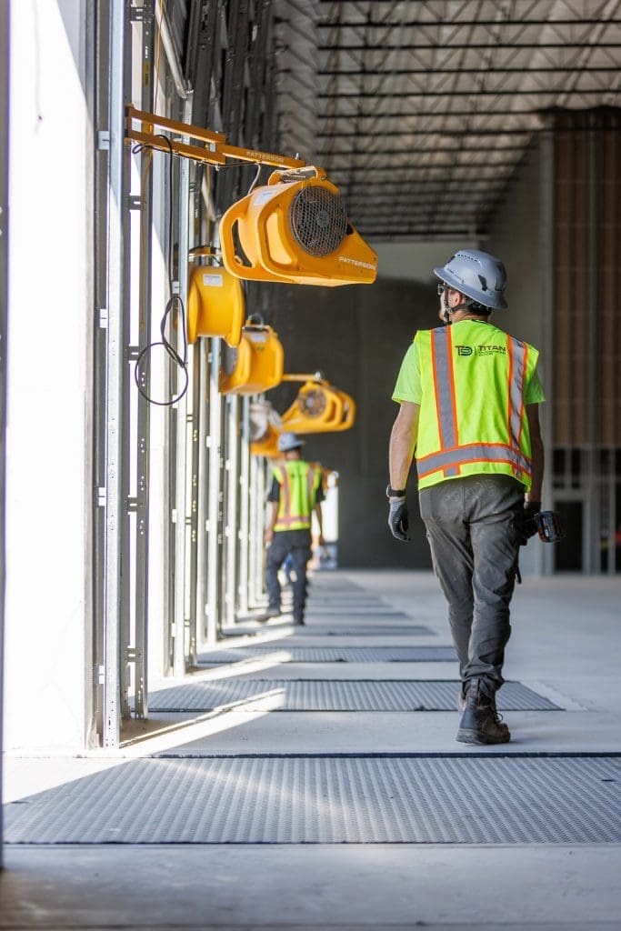 Two construction workers in safety gear walk through an industrial building with large ventilation fans mounted on the walls, showcasing the quality installations by Arizona Commercial Door Company.