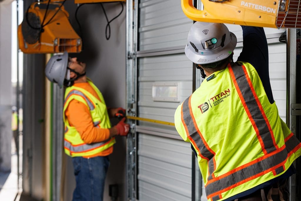 Two construction workers in safety gear install or repair a large overhead door for Arizona Commercial Door Company, using tools and measuring tape inside an industrial setting.