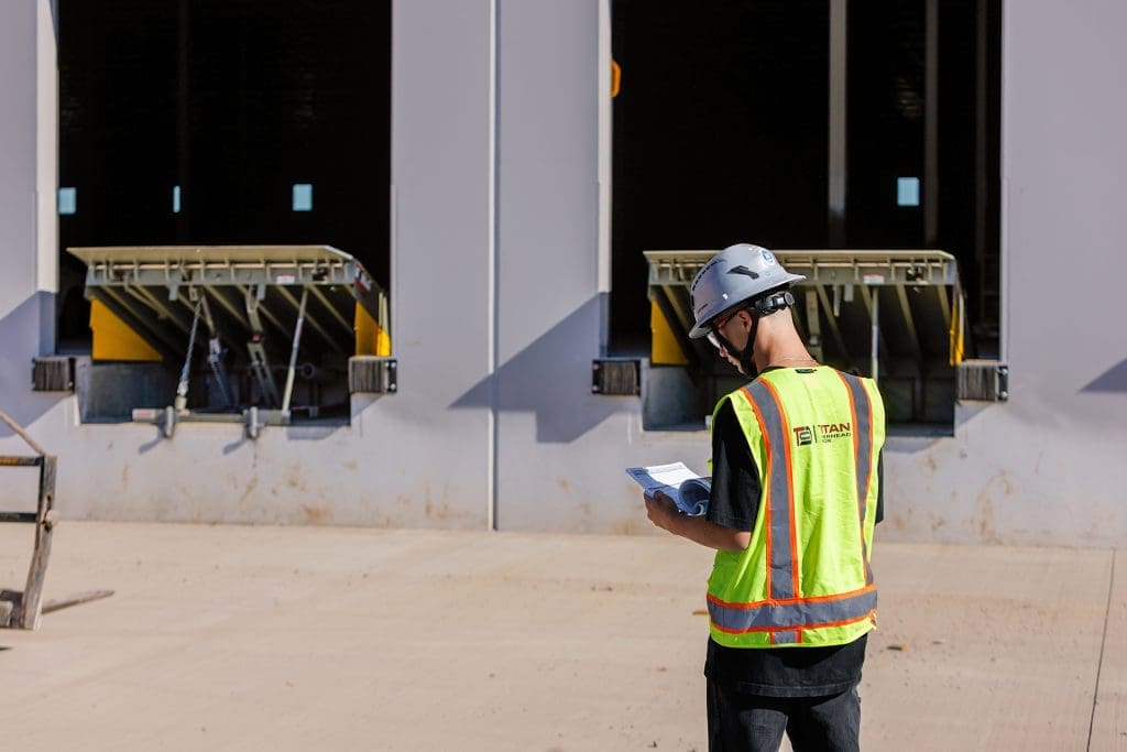 A person wearing a safety vest and helmet stands outside loading docks, holding papers and inspecting the area for proper loading dock levelers and equipment.