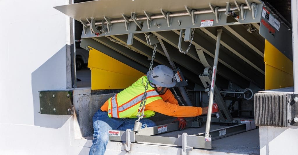 Worker in safety gear kneels and performs maintenance on a loading dock leveler, ensuring the equipment at the loading bay operates safely and efficiently.