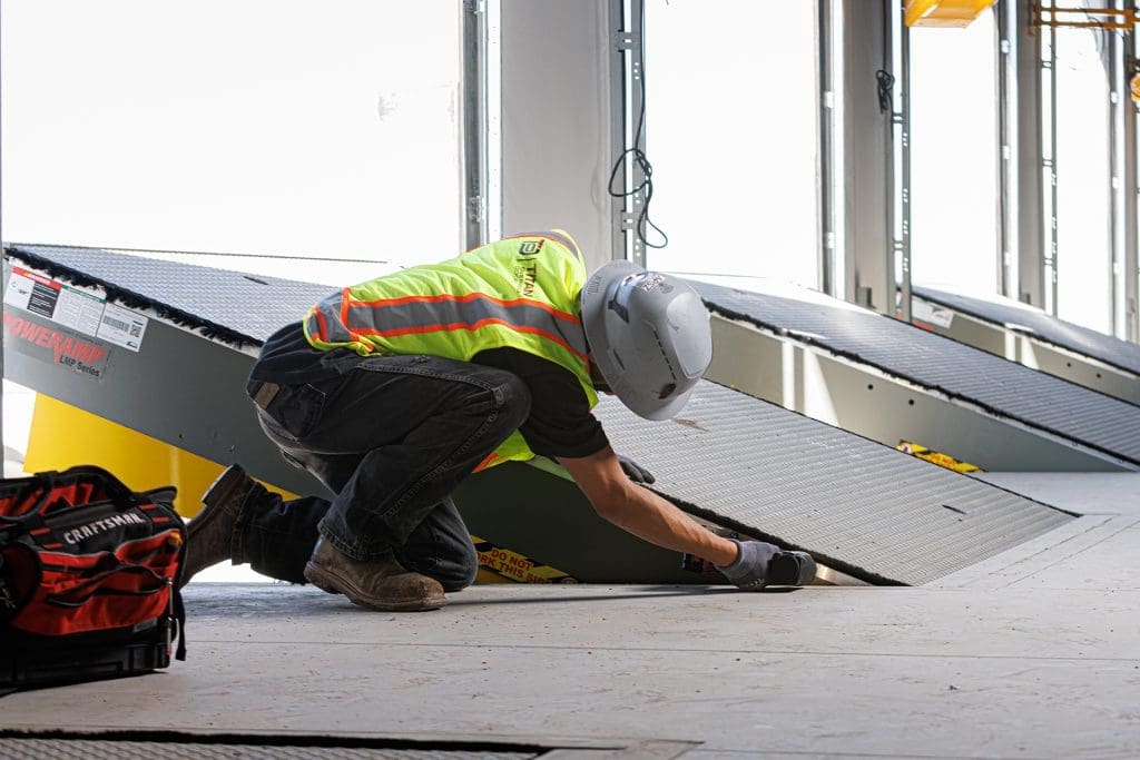 Construction worker in a safety vest and helmet installs or inspects Loading Dock Levelers and Equipment, focusing on a metal ramp at a loading dock area inside a building under construction.