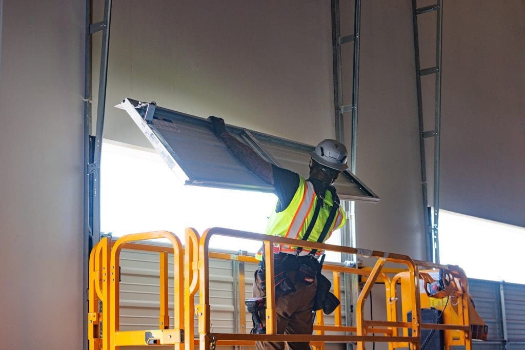 A construction worker in a safety vest and helmet installs or repairs a large overhead door while standing on a yellow mobile platform inside a building, providing 24/7 Emergency Overhead Door Repair services.