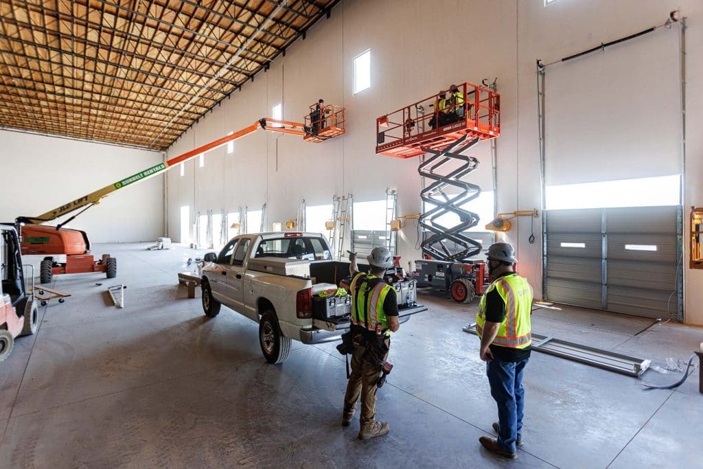 Construction workers operate lifts and stand near a pickup truck inside a large industrial warehouse with unfinished walls and ceiling, preparing the space for Commercial Dock and Door Solutions.